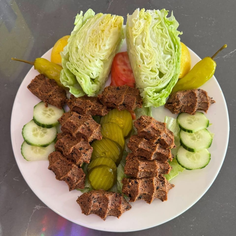 Kofte dish with lettuce, lemon, tomato, pickles, and cucumber slices on a white plate.