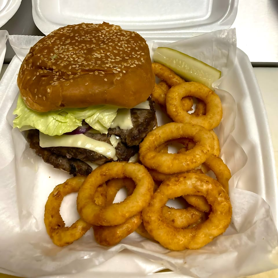 A double cheeseburger with lettuce, onion rings, and a pickle spear in a takeout container.