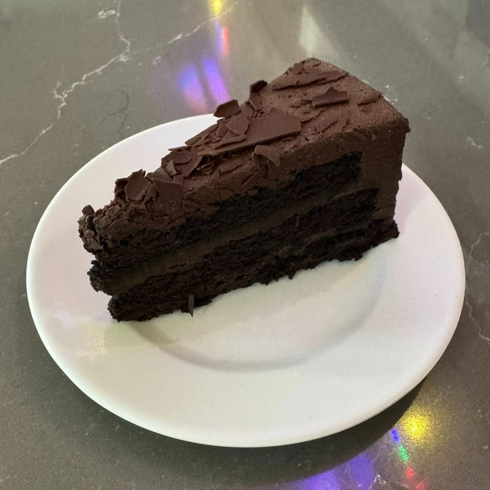 A rich slice of chocolate cake with chocolate frosting and shavings on a white plate.