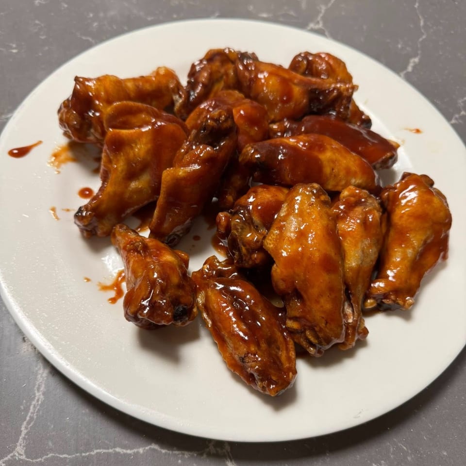 A white plate filled with Falcon Pizza's glistening, saucy chicken wings, with sauce drizzles on the plate.